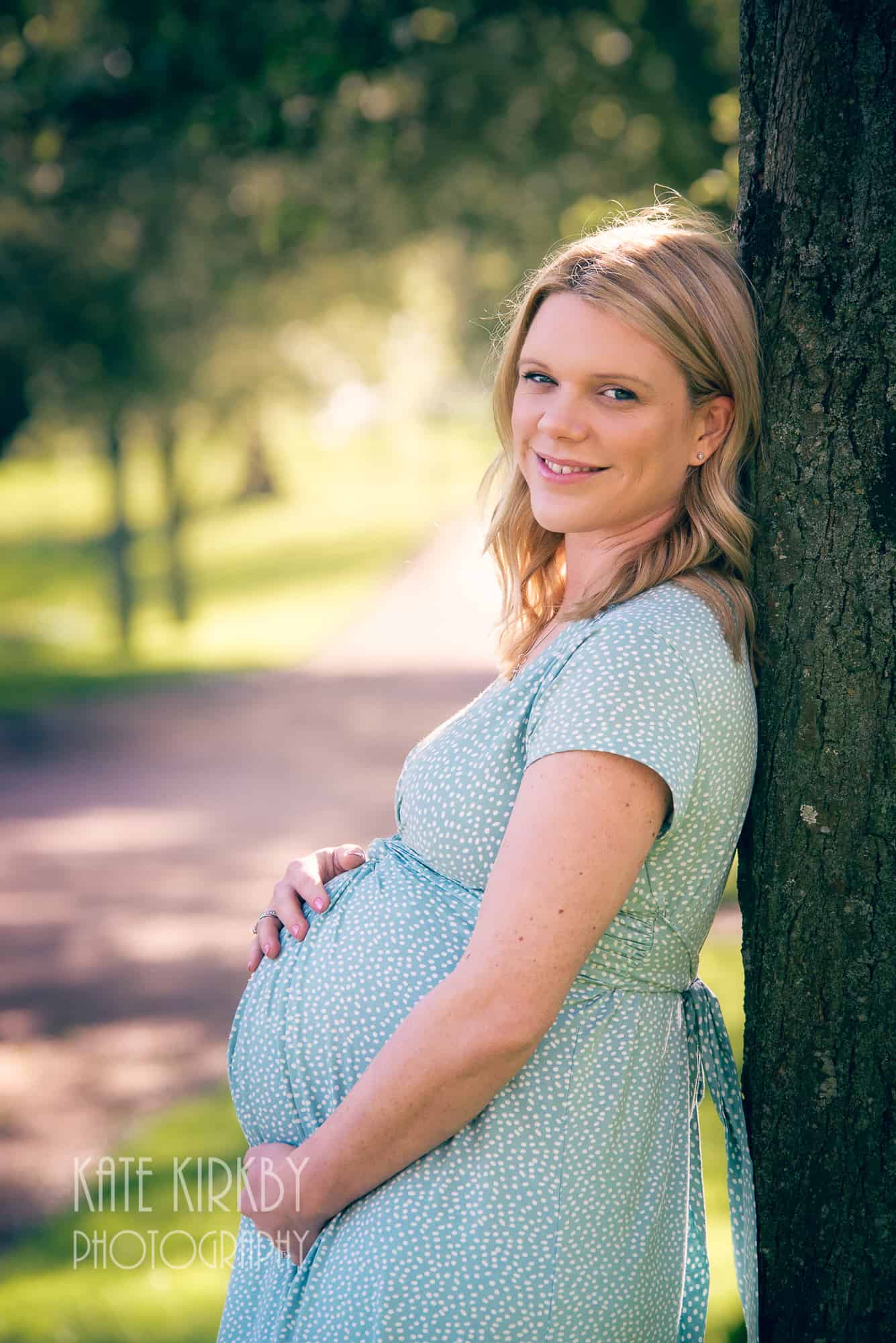 pregnant woman in green spotty dress leaning against tree