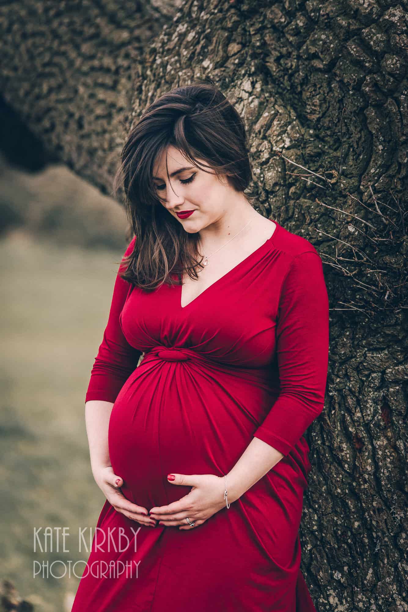 pregnant woman in red dress leaning against a tree