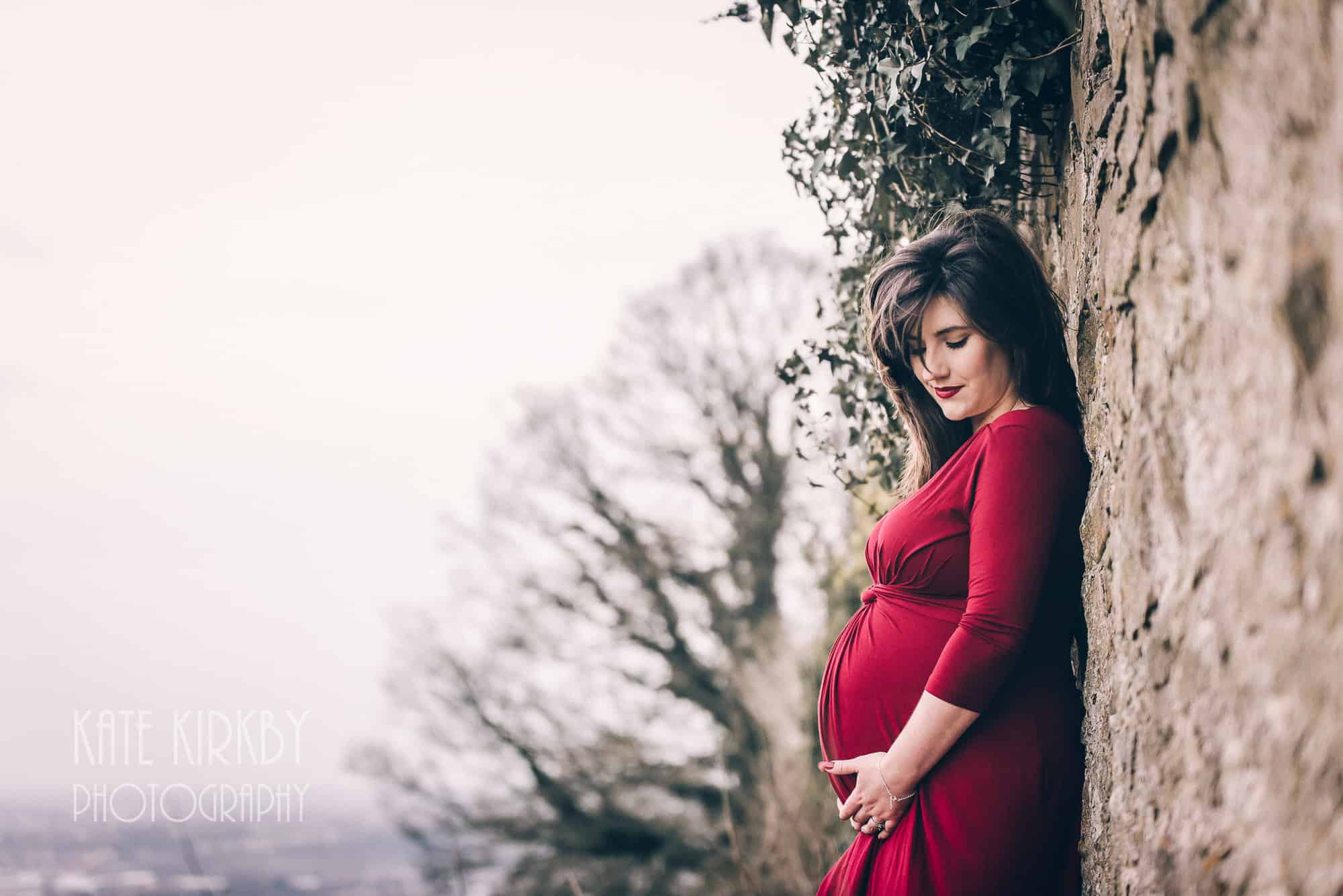 pregnant woman in red dress leaning against a wall covered in ivy