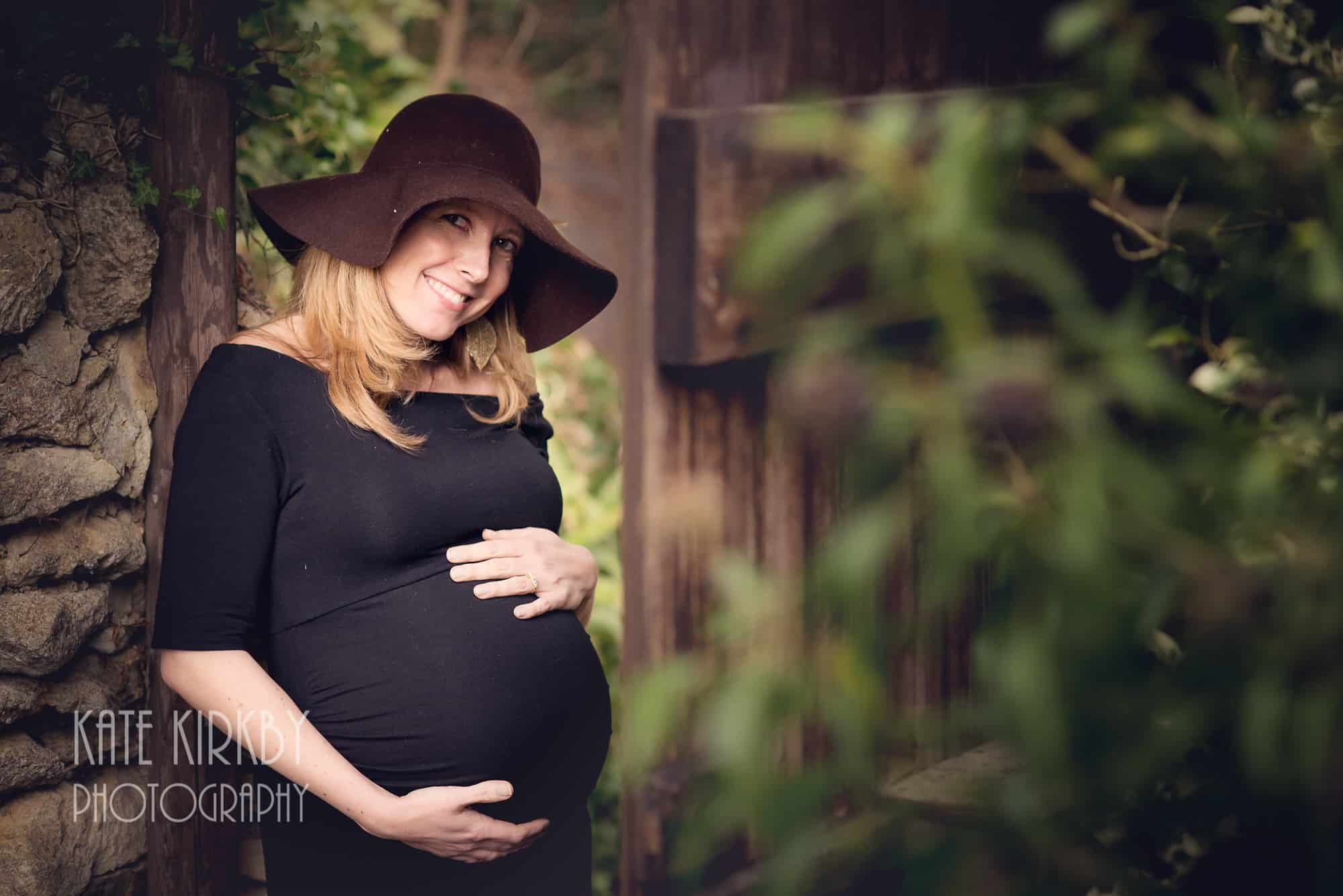 pregnant woman in black dress and brown floppy hat standing in garden gateway