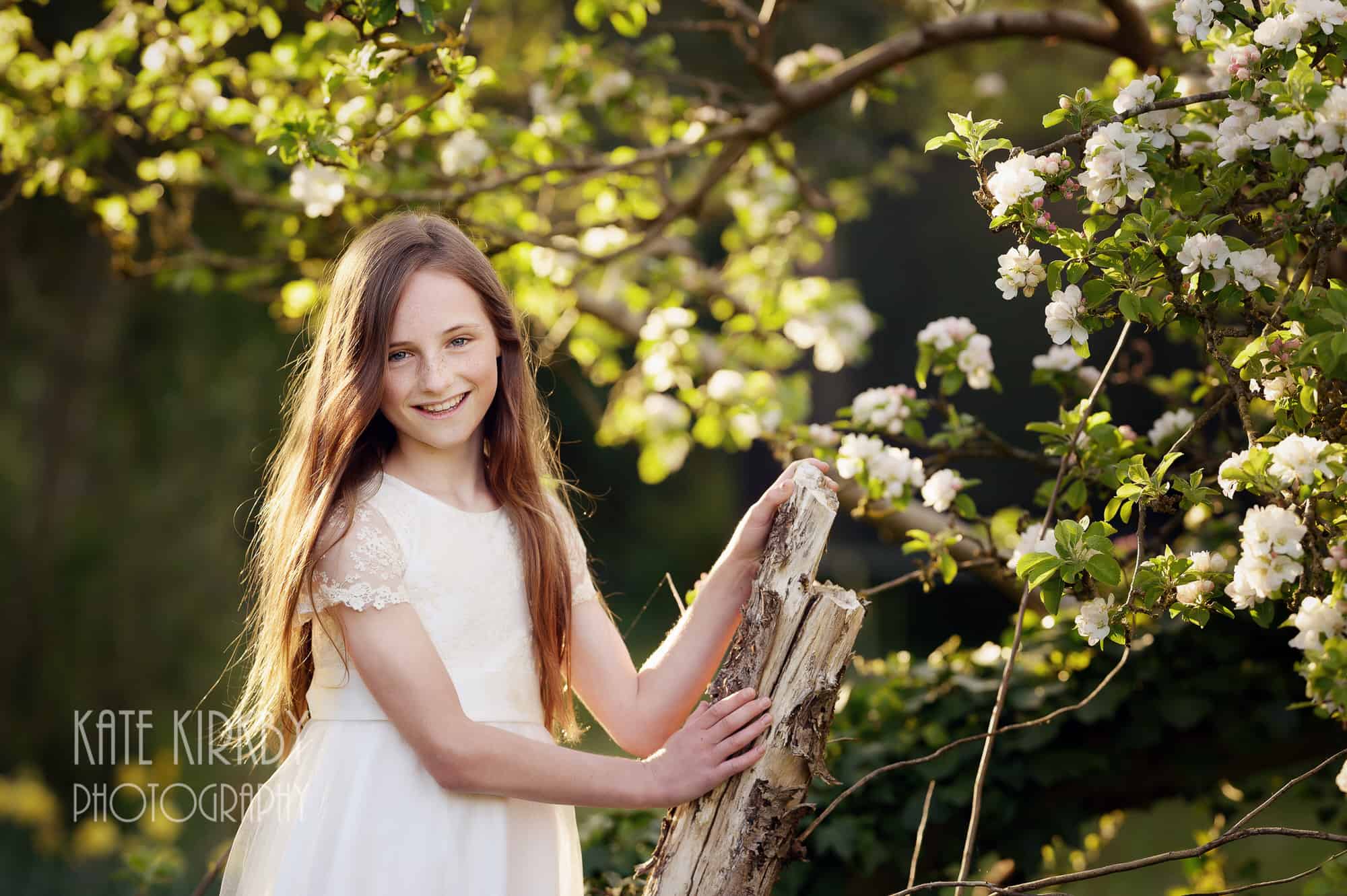girl in white dress with long drown hair standing next to tree with white blossom