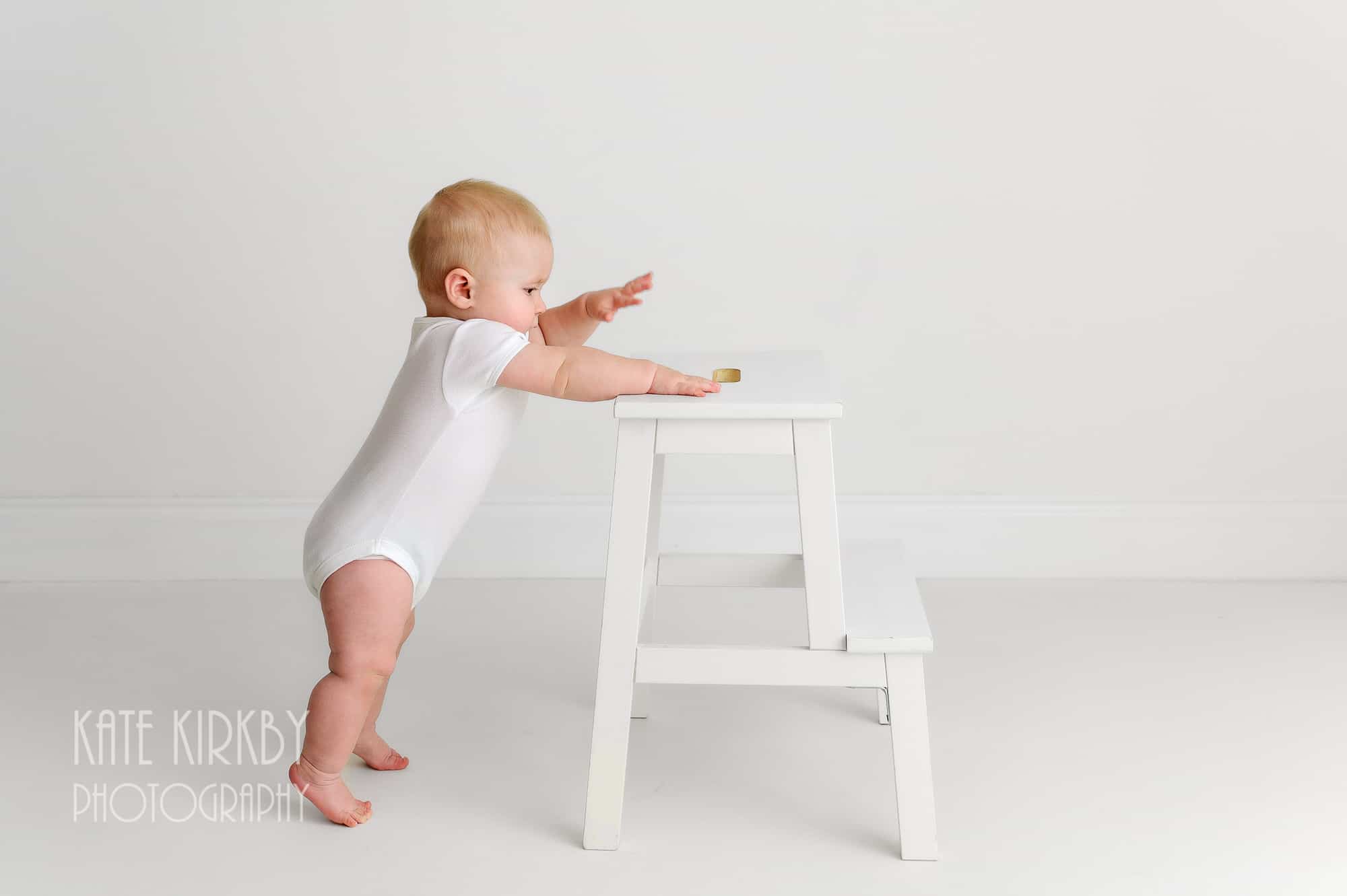 10 moth old baby in white vest holding himself up on white stool