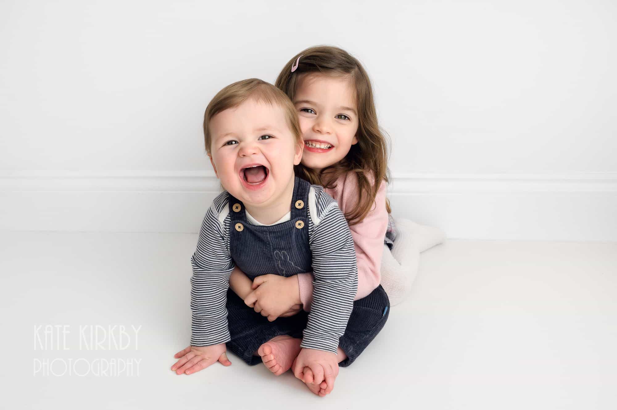 6 month old boy and his older sister cuddling and smiling at camera