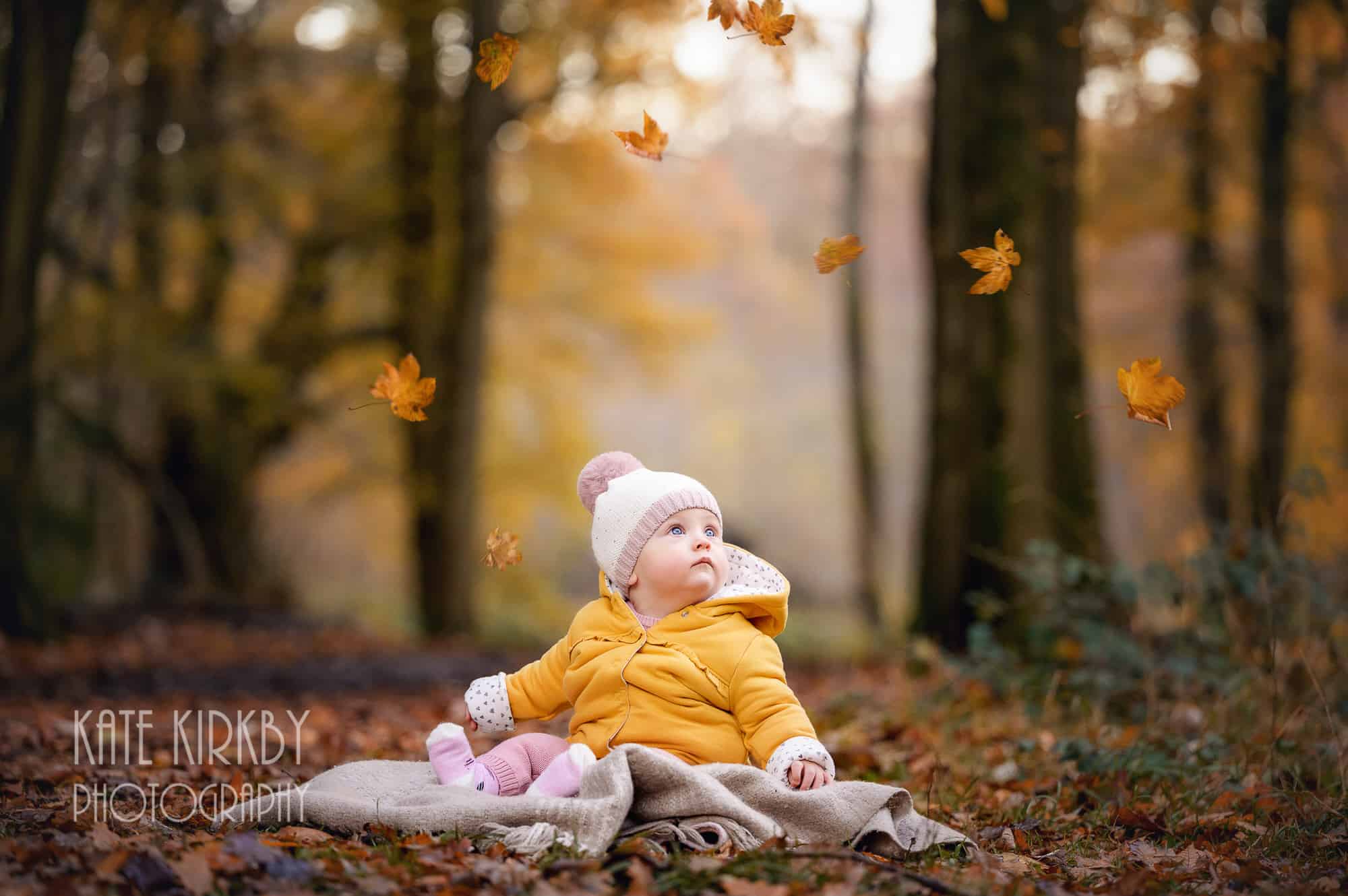 9 month old baby sitting on rug in woods with autumn leaves falling