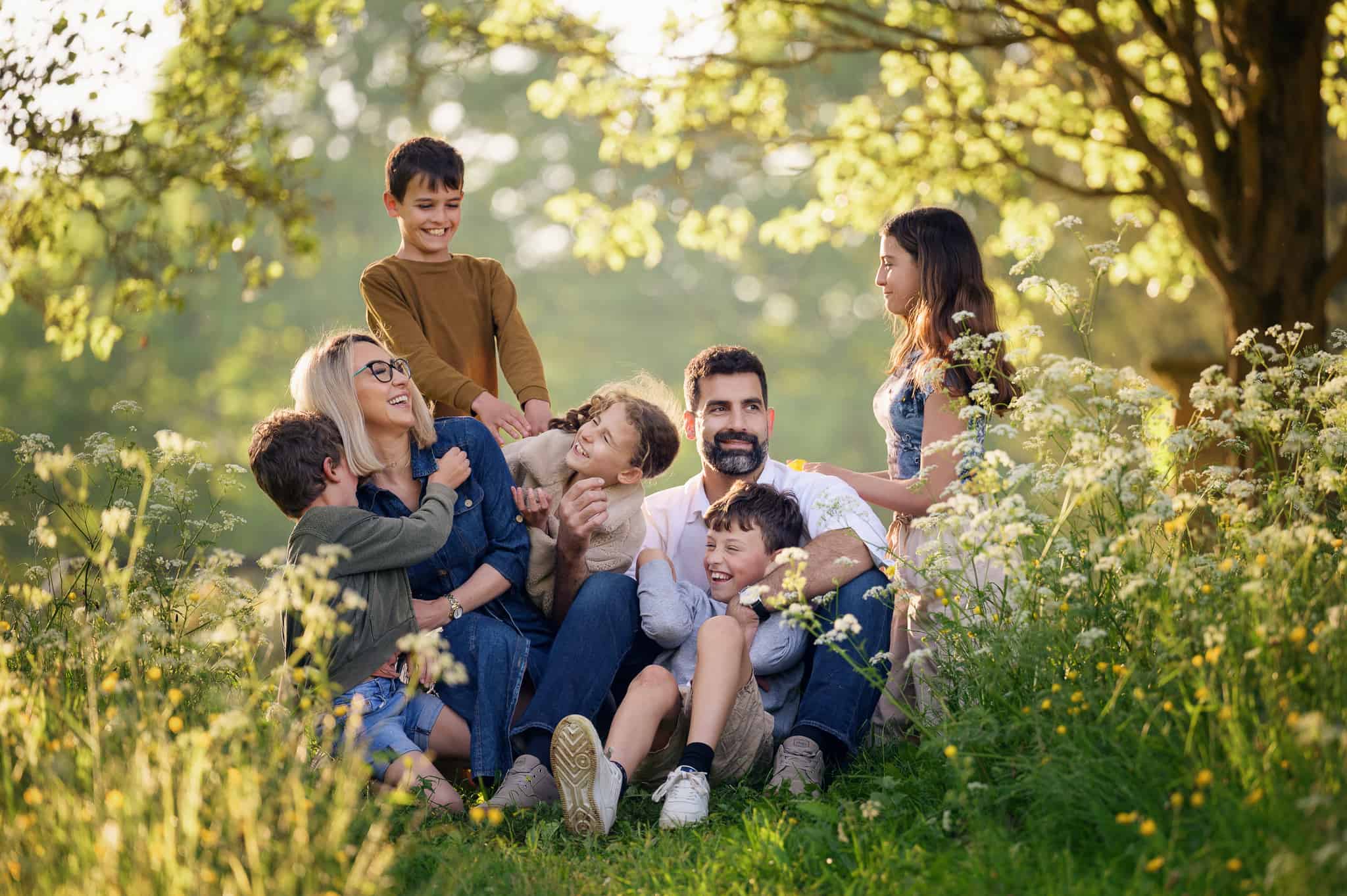 Photograph of a family sitting in a field surrounded by flowers. They are laughing and tickling each other.