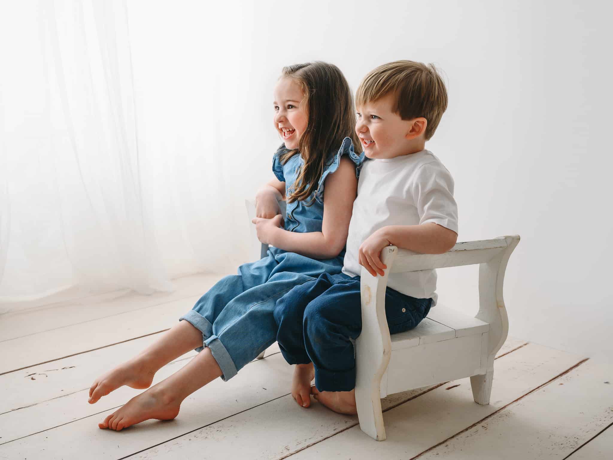 studio photograph of brother and sister sitting on a white stool looking off to the side and laughing