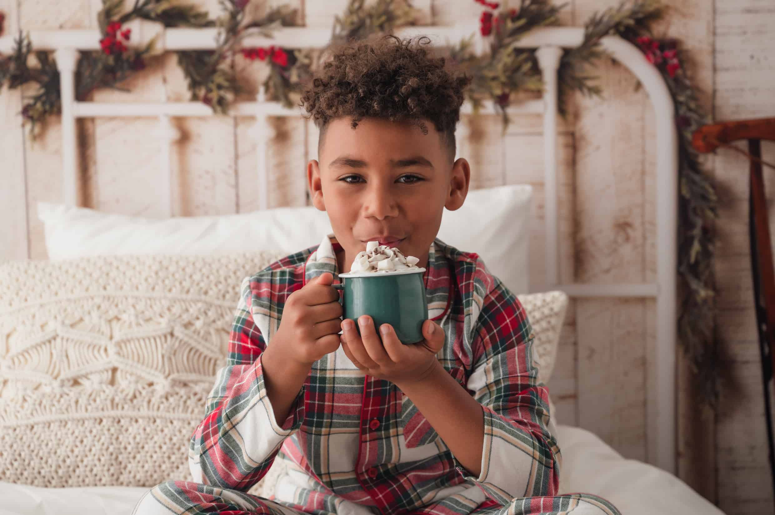 Christmas photo of a boy drinking hot chocolate sitting on a bed in tartan pyjamas