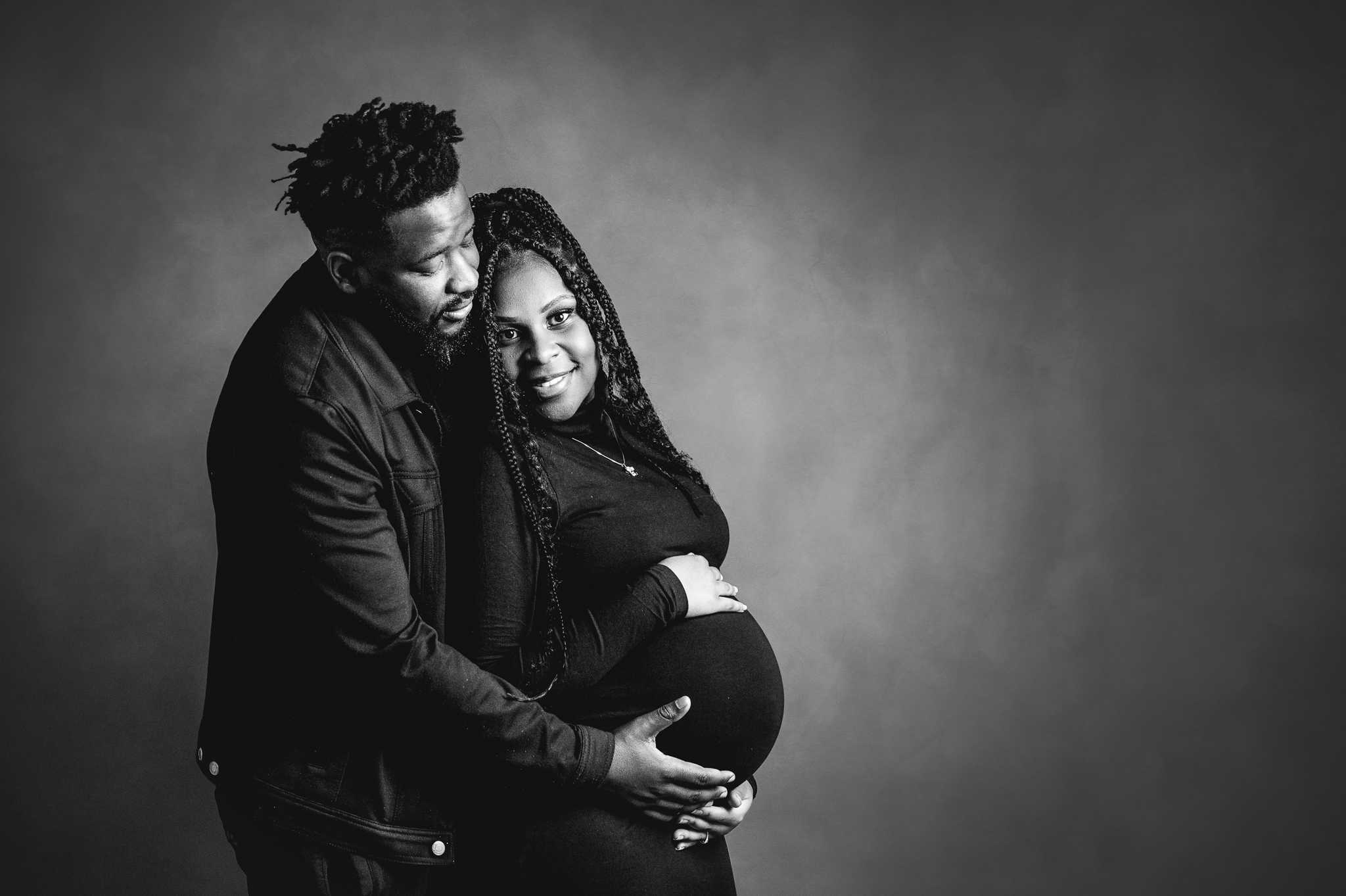 A studio photo of a couple. The lady is heavily pregnant and she is smiling at the camera